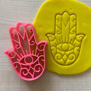 May include: A pink plastic cookie cutter in the shape of a hamsa hand with an eye in the center. The hamsa is a symbol of protection and good luck. The cookie cutter is next to a yellow piece of dough with the same hamsa design imprinted in it.