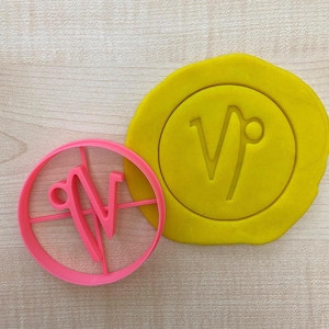 Op de afbeelding: Een roze plastic koekjesvormpje in de vorm van het astrologische teken Steenbok. De vormpje ligt op een houten oppervlak naast een geel stuk modelleerklei met hetzelfde teken erop.