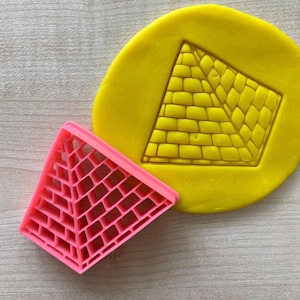Può includere: Un tagliapasta di plastica rosa a forma di piramide con un motivo a griglia. Il tagliapasta è accanto a un pezzo di plastilina gialla con un'impressione a forma di piramide.