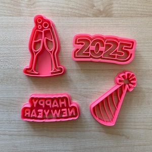 May include: Four pink plastic cookie cutters for New Year's Eve. The cutters are shaped like a champagne flute, a party hat, the year 2025, and the words "Happy New Year".