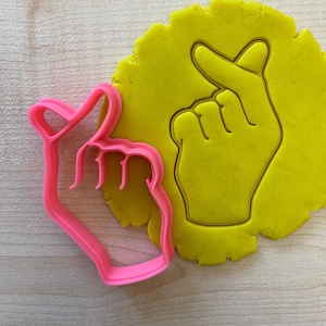 Può includere: Un tagliabiscotti in plastica rosa a forma di mano che fa un cuore, accanto a un pezzo di pasta gialla con la stessa forma di mano impressa. Il tagliabiscotti e la pasta sono su una superficie di legno chiaro.