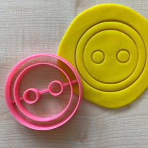 Può includere: Due stampini per biscotti in plastica, uno giallo e uno rosa, a forma di bottoni con due fori. Lo stampino giallo è più grande dello stampino rosa.