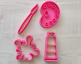 May include: Four pink plastic cookie cutters shaped like a paintbrush, paint palette, paint tube, and a paint splatter.