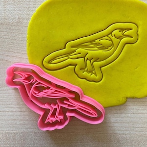 Può includere: Un tagliapasta in plastica rosa a forma di uccello. Il tagliapasta è mostrato accanto a un pezzo di plastilina gialla, che ha l'impressione dell'uccello.