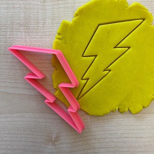 Può includere: Un tagliapasta a forma di fulmine in plastica rosa con un'impronta di fulmine gialla in plastilina.