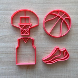 May include: Four pink plastic cookie cutters shaped like a basketball, a basketball hoop, a basketball jersey, and a sneaker.