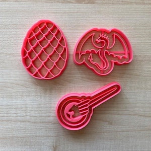 Peut inclure: Trois emportes-pièces en plastique rose en forme de dragon, d'œuf de dragon et d'une main tenant un œuf de dragon.