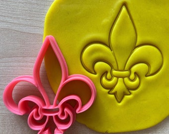 Könnte beinhalten: Ein rosa Plastik-Ausstecher in Form einer Fleur-de-lis. Der Ausstecher liegt auf einem gelben Fondantblatt mit einer Abformung einer Fleur-de-lis.