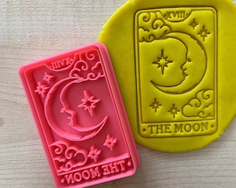 May include: Pink plastic cookie cutter shaped like a tarot card with the image of a crescent moon and stars. The card reads "THE MOON" in a stylized font.