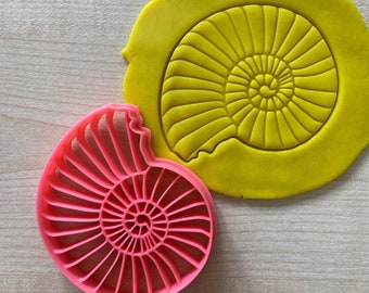 May include: A pink plastic cookie cutter shaped like an ammonite, a spiral-shaped fossil. The cutter is shown next to a yellow piece of modeling clay that has been pressed with the cutter to create an impression.