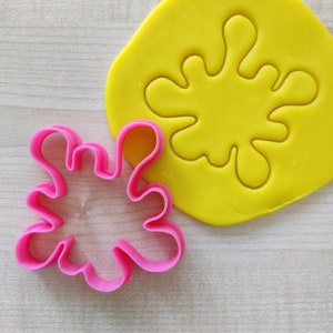 Può includere: Un tagliabiscotti rosa a forma di schizzo in plastica accanto a un pezzo di plastilina gialla con lo stesso disegno a schizzo premuto. Il tagliabiscotti ha un contorno ondulato e la plastilina è liscia. Lo sfondo è un legno chiaro.