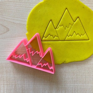 May include: Pink plastic cookie cutter shaped like three mountains with a wavy top. The cutter is next to a yellow piece of modelling clay with the same mountain design imprinted on it.