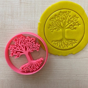 Może przedstawiać: Różowa plastikowa foremka do ciasteczek w kształcie drzewa z korzeniami. Foremka leży na drewnianej powierzchni obok żółtego kawałka gliny z tym samym wzorem drzewa.