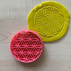 May include: A pink plastic cookie cutter shaped like a flower of life design. The cutter is on a wooden surface next to a yellow piece of dough with the same design imprinted on it.