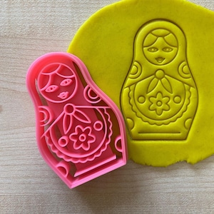 Peut inclure: Emporte-pièce en plastique rose en forme de poupée russe traditionnelle, ou poupée matriochka, avec des motifs floraux complexes. L'emporte-pièce est présenté sur de l'argile à modeler jaune, avec l'empreinte de l'emporte-pièce visible dans l'argile.
