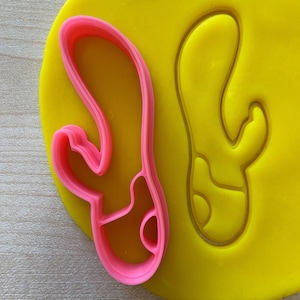 May include: A pink plastic cookie cutter shaped like a cactus with a single arm. The cutter is on top of a yellow plastic surface.