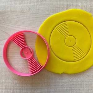 May include: A pink plastic cookie cutter shaped like a record with a yellow fondant cutout of a record.
