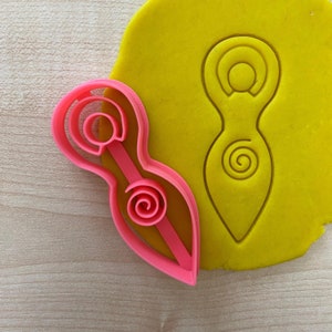 May include: Pink plastic cookie cutter shaped like a female figure with a spiral design. The cutter is on a yellow surface.