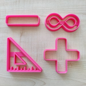 Puede incluir: Conjunto de cuatro cortadores de galletas de plástico rosa en forma de rectángulo, símbolo de infinito, signo más y transportador.