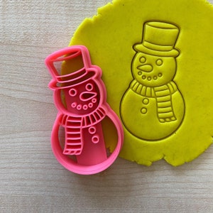 May include: Pink plastic snowman cookie cutter with a red and white striped scarf. The cutter is on top of a yellow play dough snowman.
