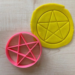 May include: A pink plastic cookie cutter shaped like a five-pointed star with a yellow circle of dough behind it.