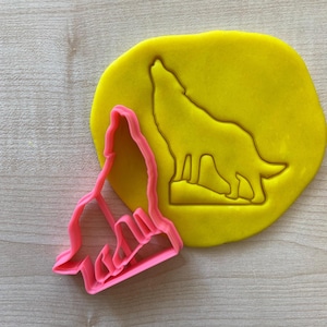 Peut inclure: Un emporte-pièce en plastique rose en forme de loup hurlant, à côté d'un morceau de pâte jaune avec le contour du loup pressé dedans. Le loup hurle, la tête en arrière.