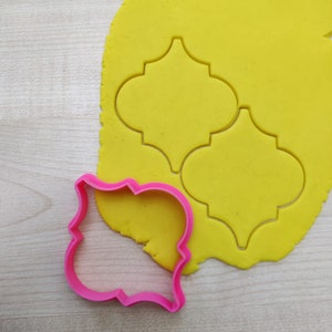 May include: A pink plastic cookie cutter in the shape of a decorative frame, next to a yellow sheet of rolled out cookie dough with two cut-out shapes of a Moroccan tile design.