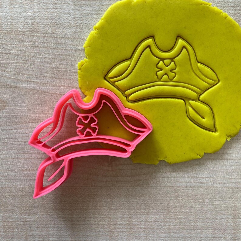 May include: Pink plastic pirate hat cookie cutter with a red cross design. The cutter is shown on top of a yellow piece of cookie dough.