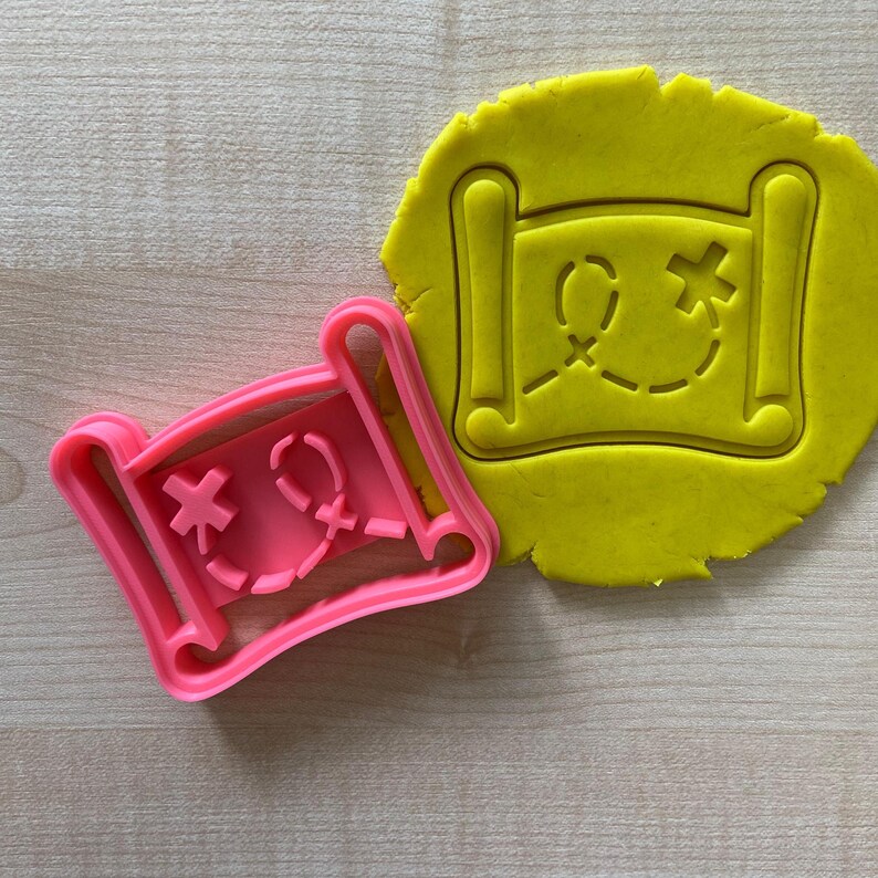 May include: Pink plastic cookie cutter shaped like a rolled up treasure map with an X marking the spot. The cutter is on top of a yellow piece of rolled out cookie dough.