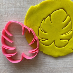 May include: A pink plastic cookie cutter shaped like a monstera leaf, next to a yellow piece of modeling clay with the same leaf shape cut out.