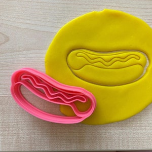 Może przedstawiać: Różowa plastikowa foremka do ciasteczek w kształcie hot doga z falistymi liniami. Foremka leży na żółtym kawałku plasteliny z wyciętym kształtem hot doga.