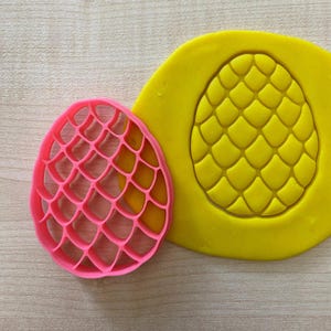 May include: A pink plastic cookie cutter shaped like a pineapple with a textured surface. A yellow piece of modeling clay is imprinted with the pineapple design.