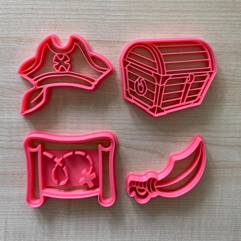 May include: Set of four pink plastic cookie cutters shaped like a pirate hat, a treasure chest, a map, and a sword.