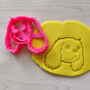 May include: Pink plastic cookie cutter shaped like a Cavalier King Charles Spaniel dog, with a yellow fondant cookie being cut out with the cutter.