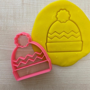 May include: Pink plastic cookie cutter shaped like a winter hat with a pom-pom on top. The hat has a chevron pattern.