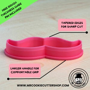 May include: Pink plastic moustache cookie cutter with a larger handle for a comfortable grip. The cutter has tapered edges for a sharp cut. The text "FREE DOUGH THICKNESS GUIDE PIN INCLUDED" is in the top left corner of the image. The text "TAPERED EDGES FOR SHARP CUT" is in the top right corner of the image. The text "LARGER HANDLE FOR COMFORTABLE GRIP" is in the bottom left corner of the image. The text "WWW.MRCOOKIECUTTERSHOP.COM" is in the bottom centre of the image. The text "Mr. CookieCutter" is in the bottom right corner of the image.