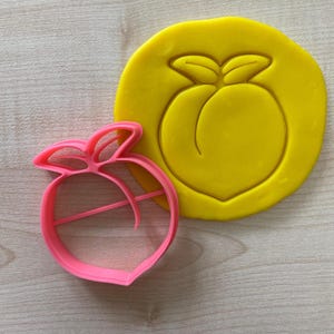 May include: A pink plastic peach-shaped cookie cutter with a yellow peach imprint on a yellow fondant background.