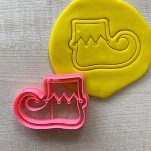 May include: Pink plastic cookie cutter shaped like an elf shoe. The cutter is on a yellow surface.