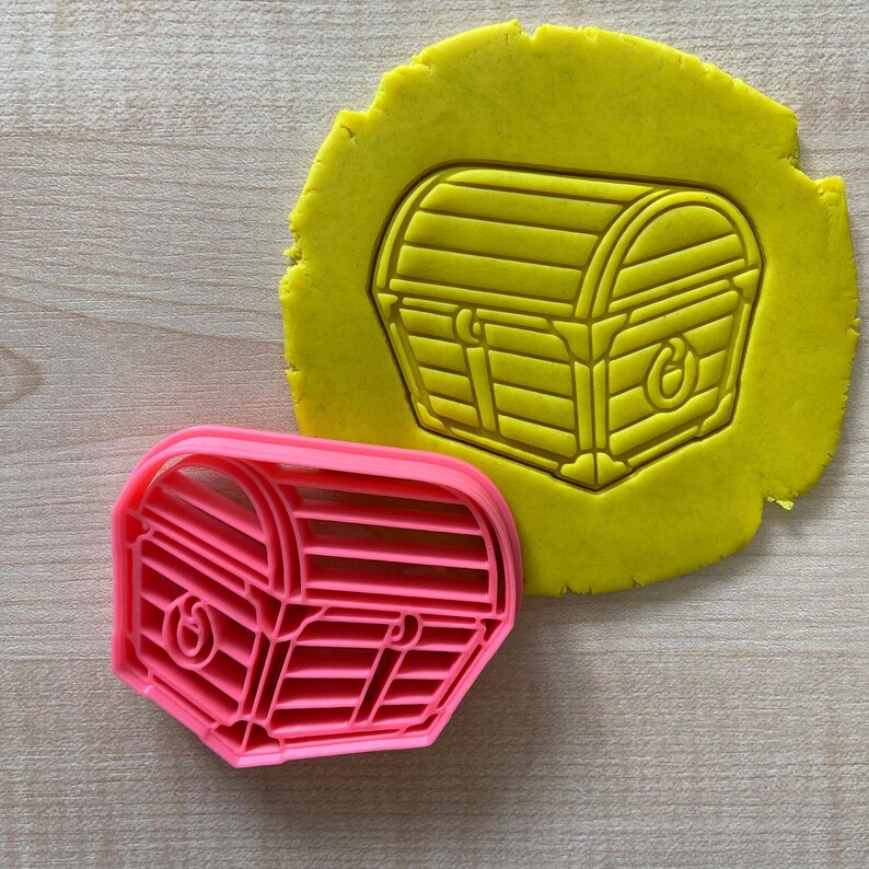 May include: A pink plastic cookie cutter shaped like a treasure chest with a hinged lid and a lock. The cutter is shown on top of a yellow piece of cookie dough.