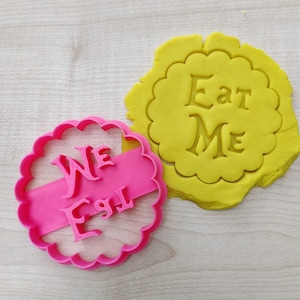 Op de afbeelding: Twee roze plastic koekjesvormen, een met de woorden "We Eat" en de andere met de woorden "Eat Me".