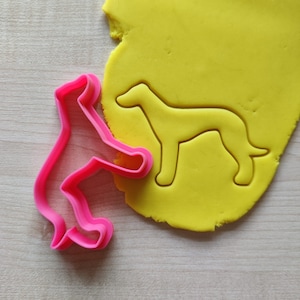 May include: Pink plastic cookie cutter shaped like a dog, next to a yellow piece of dough with a dog-shaped impression.
