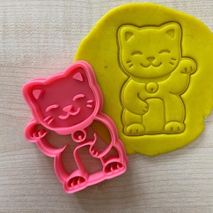 Może przedstawiać: Różowa plastikowa foremka do ciasteczek w kształcie japońskiego Maneki-Neko, czyli kota szczęścia, z uniesioną łapą. Foremka do ciasteczek znajduje się obok odcisku żółtego ciasta do ciasteczek o tym samym wzorze.