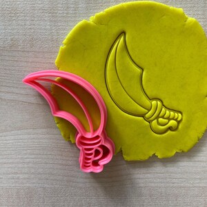 May include: Pink plastic cookie cutter shaped like a curved sword with a decorative handle. The cutter is on top of yellow play dough with an impression of the sword.
