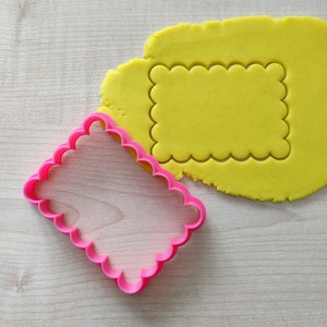 Può includere: Un tagliapasta di plastica rosa a forma di rettangolo con bordi a festoni. Il tagliapasta è sopra un pezzo di pasta per biscotti gialla che è stata tagliata con la stessa forma.