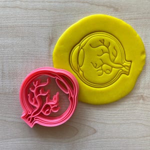 May include: A pink plastic cookie cutter shaped like a human eye. The cutter is layered to create a detailed design. The cutter is shown on a yellow surface.