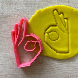 May include: A pink plastic cookie cutter in the shape of the "okay" hand gesture, next to a yellow playdough imprint of the same shape. The cutter is on a light wood surface.