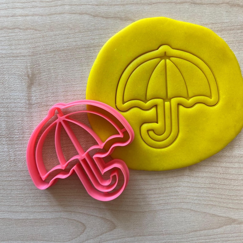 Puede incluir: Un cortador de galletas de pl&aacute;stico rosa con forma de paraguas. El cortador est&aacute; sobre una superficie amarilla.