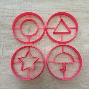 Pode incluir: Quatro cortadores de biscoitos de plástico rosa em forma de círculo, triângulo, estrela e guarda-chuva.