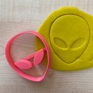 May include: Pink plastic cookie cutter shaped like an alien head with large eyes. The cutter is shown next to a yellow piece of dough with the alien head design pressed into it.