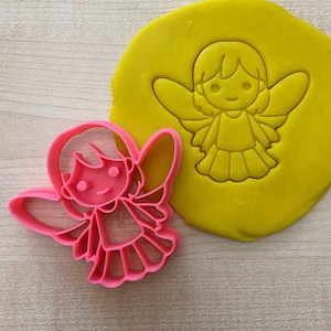 May include: Pink plastic cookie cutter shaped like an angel with wings, a dress, and a smiling face. The cutter is shown next to a yellow imprint of the cutter in dough.